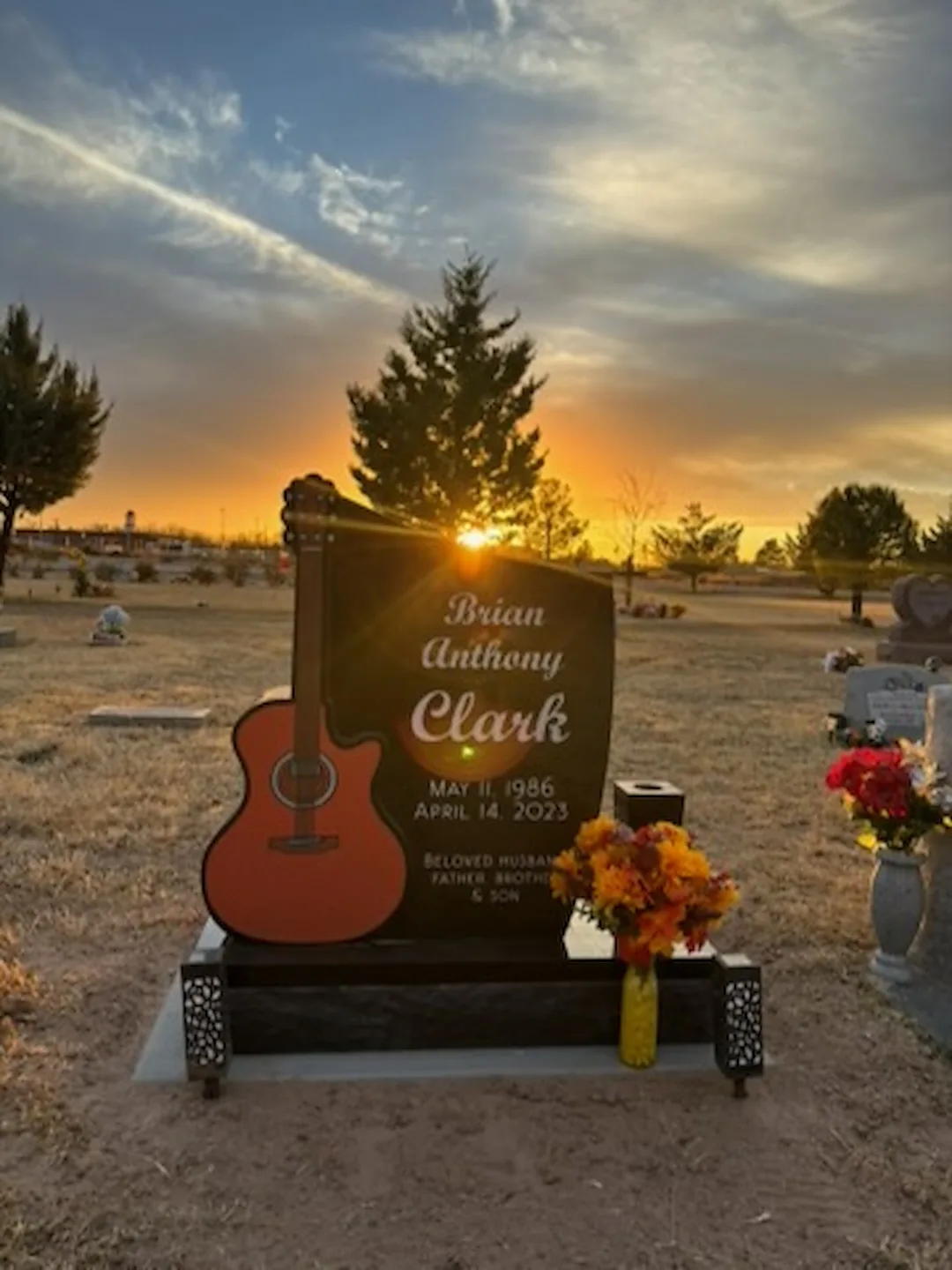 Custom guitar-shaped black granite headstone at sunset in a Las Cruces, New Mexico cemetery, with a red acoustic guitar inlay and hand-pierced scrollwork base, crafted by Rose Rock Monument